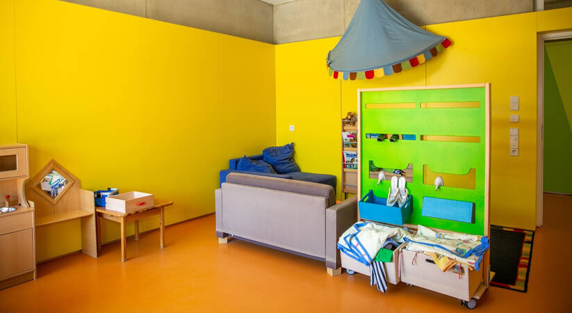 Ein helles, kindgerecht eingerichtetes Zimmer mit gelben Wänden, einem Sofa, einer Spielecke und bunten Dekorationen.