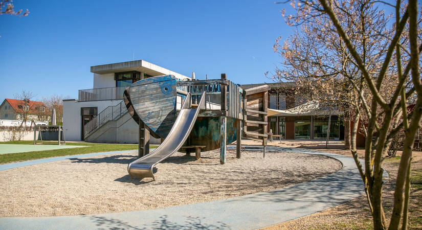 Ein Spielplatz mit einem großen hölzernen Schiffsrumpf und einer Metallrutsche befindet sich vor einem modernen Gebäude.
