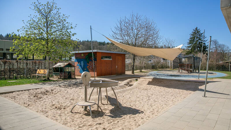 Ein Spielplatz mit Sandkasten, Kletterstruktur und Sonnensegel, umgeben von Bäumen und einem gemauerten Nebengebäude.