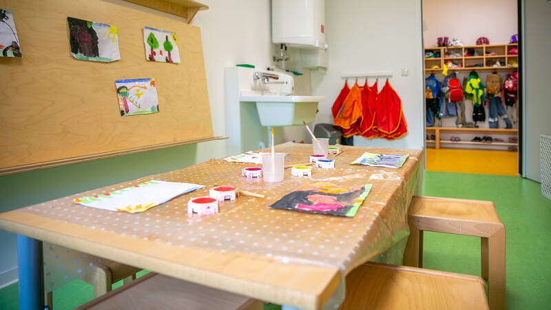 Ein heller Kindergartenraum mit einem Tisch voller Malutensilien, Kinderzeichnungen an der Wand, farbigen Haken mit Jacken und einem Regal voller Kleidung und Schuhe im Hintergrund.