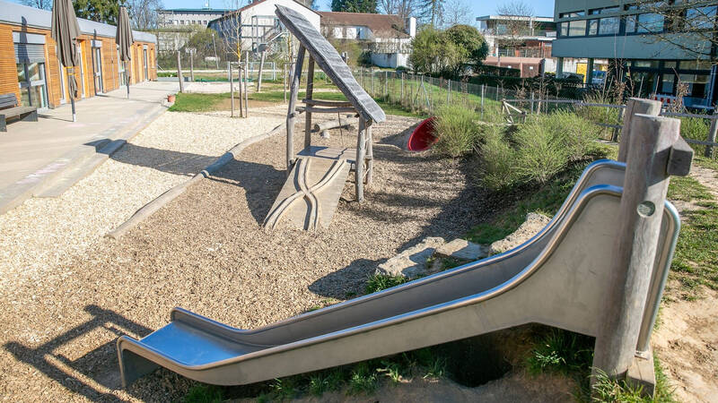 Ein Spielplatz mit Rutsche, Holzklettergerüst und Holzspänen am Boden, umgeben von Gebäuden und Büschen.