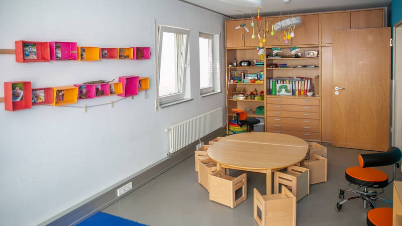 Ein heller Kinderzimmerraum mit einem runden Holztisch, umgeben von kleinen Stühlen, bunten Regalen an der Wand und einem großen Holzschrank mit Spielzeug und Büchern.