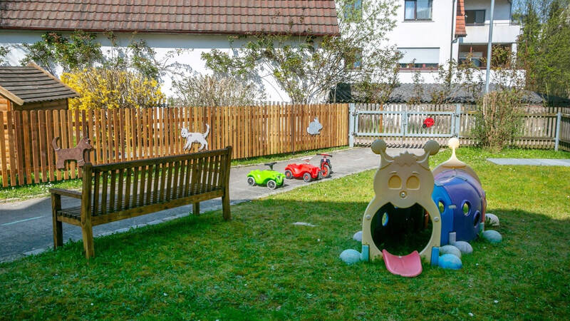 Ein Spielplatz mit einer Holzbank, einem bunten Krabbeltunnel in Form eines Tieres und kleinen Spielzeugautos auf einer Grasfläche, umgeben von einem Holzzaun.