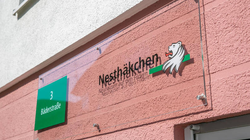 Ein Schild an einer rosa Wand zeigt den Straßennamen "Bäderstraße 3" und das Logo einer Einrichtung namens "Nesthäkchen" mit einem stilisierten Löwenkopf.
