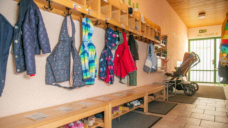In einem Flur befinden sich an einer Seite bunte Kinderjacken auf Haken, darunter Bänke mit Schuhen, und am Ende steht ein Kinderwagen nahe einer Tür.