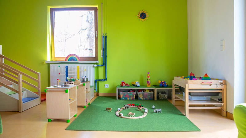 Ein bunter Kindergartenraum mit Spielzeug, Kindermöbeln und einem grünen Teppich, umgeben von einer hellgrünen Wand.