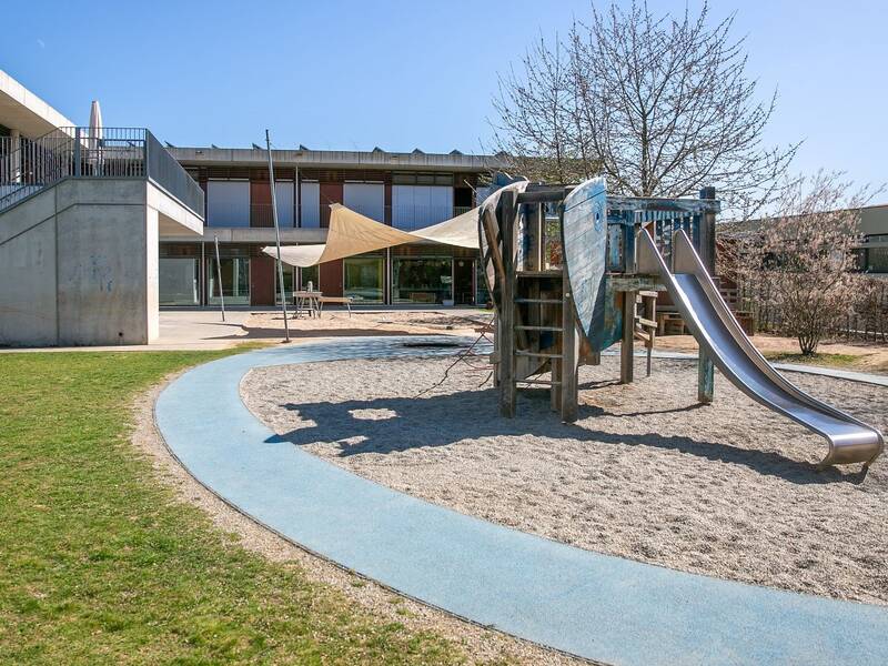 Der Spielplatz auf dem Bild zeigt eine Rutsche und Kletterelemente, umgeben von einem gepflasterten Weg und einer Wiese, mit einem modernen Gebäude im Hintergrund.