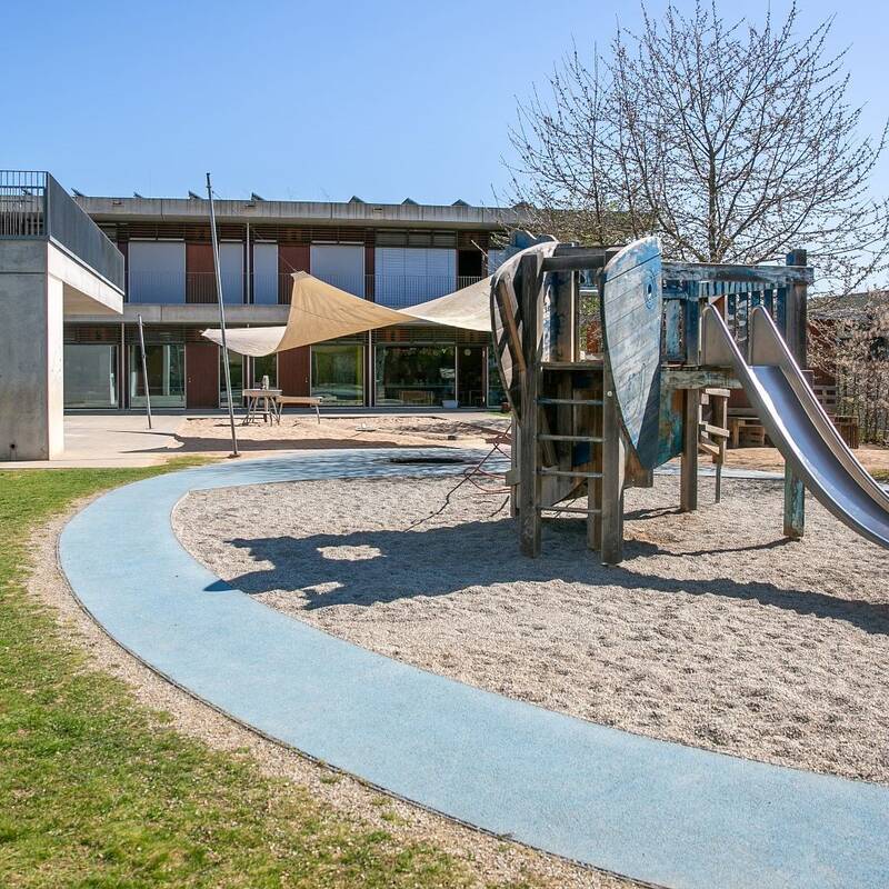 Der Spielplatz auf dem Bild zeigt eine Rutsche und Kletterelemente, umgeben von einem gepflasterten Weg und einer Wiese, mit einem modernen Gebäude im Hintergrund.